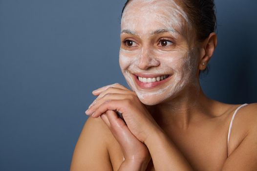 Attractive woman smiles with beautiful toothy smile, posing against gray background with facial mask, removing make-up and cleaning her face using exfoliant facial cleansing beauty cosmetic product