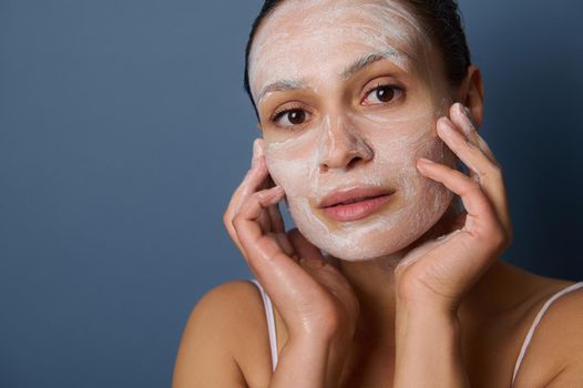 Close-up of a young pretty dark-skinned woman giving herself face massage, removing make-up and cleaning her face using exfoliant facial cleansing beauty cosmetic product, isolated on gray background