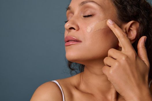 Close-up of attractive woman with her eyes closed applying liquid tonal foundation fluid on her face with her finger, posing against gray wall background with copy space. Make-up, skin care concept