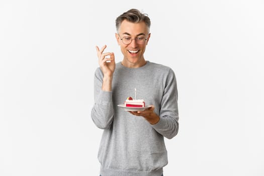 Portrait of handsome middle-aged man, smiling and looking amazed at birthday cake with lit candle, making wish and celebrating b-day, standing over white background.