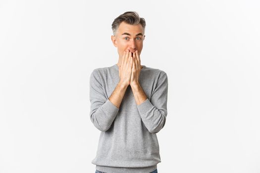 Image of excited and amused caucasian man with gray hair, cover mouth and looking at something interesting, standing over white background.