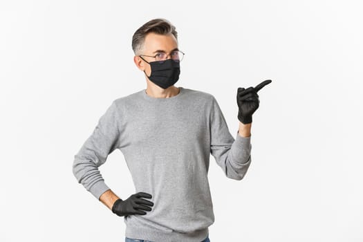 Concept of coronavirus, lifestyle and quarantine. Image of middle-aged man in glasses, medical mask and gloves, pointing finger right, showing promo banner, standing over white background.