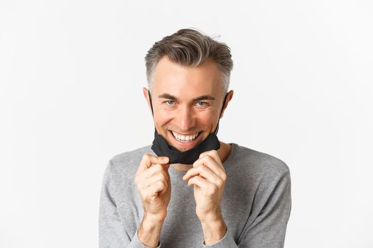 Close-up of handsome middle-aged man, taking-off medical mask and looking happy, smiling pleased, standing over white background. Concept of covid-19, social distancing and quarantine.