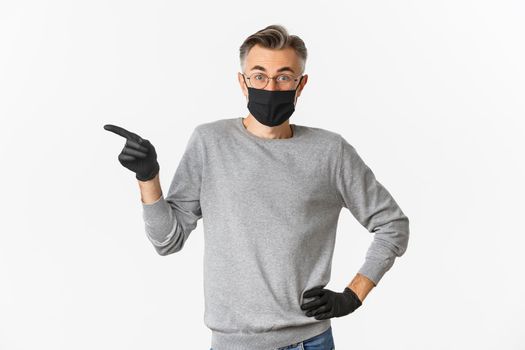 Concept of coronavirus, lifestyle and quarantine. Portrait of handsome middle-aged man in glasses, medical mask and gloves, asking question with curious face, pointing finger left, white background.