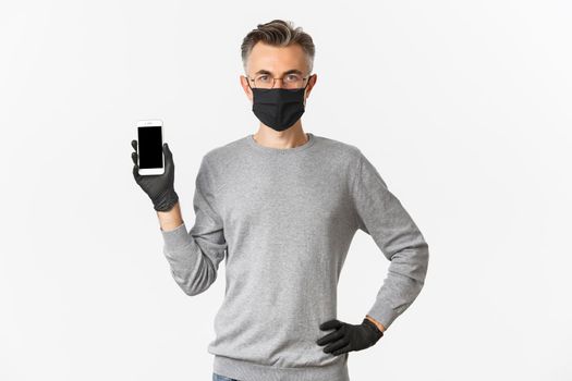 Concept of covid-19, social distancing and lifestyle. Image of confident middle-aged man in glasses, medical mask and gloves, showing smartphone screen, demonstrate an app.