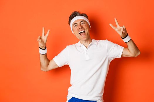 Image of happy and healthy middle-aged man in sport clothing, showing peace signs and smiling, workout over orange background.