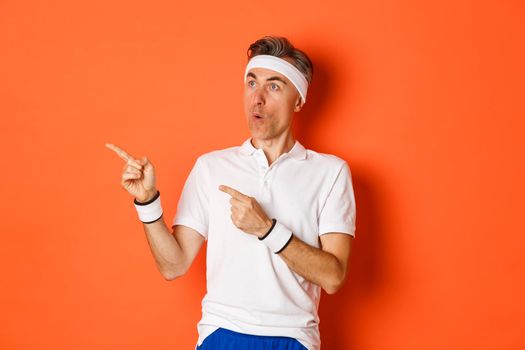 Image of intrigued middle-aged sportsman in activewear, pointing and looking left at advertisement about workout or sports, standing over orange background.