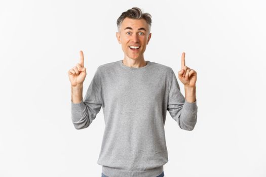 Image of amazed adult guy with grey short hairstyle, showing awesome offer, pointing fingers up at copy space and smiling happy, standing over white background.