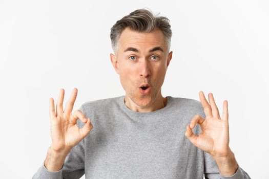 Close-up of impressed middle-aged guy, showing okay signs in approval, like something good, recommending product, standing over white background.