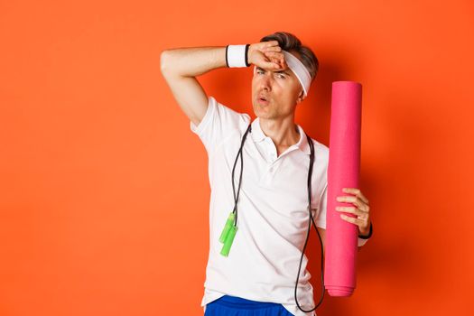 Concept of workout, gym and lifestyle. Image of handsome middle-aged man, tired after fitness exercises, holding skipping rope and yoga mat, wiping sweat off forehead, orange background.