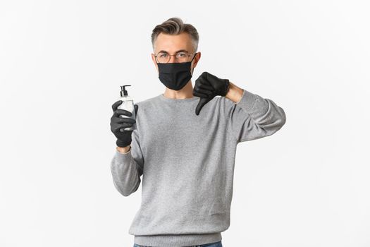 Concept of coronavirus, lifestyle and quarantine. Portrait of middle-aged man in medical mask and gloves do not recommend antiseptic, showing hand sanitizer and thumbs-down, white background.