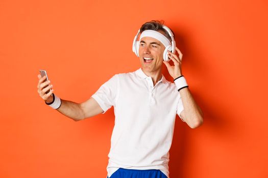 Portrait of handsome middle-aged male athlete, wearing gym uniform, listening music in headphones and taking selfie on mobile phone during workout, standing over orange background.