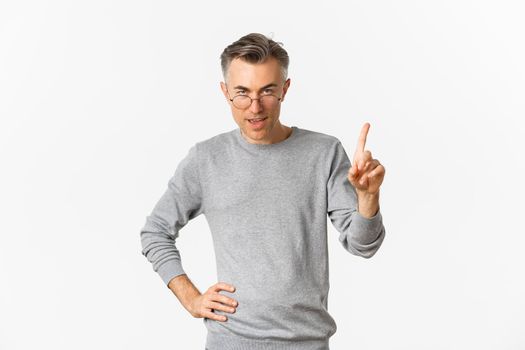 Image of handsome middle-aged man in glasses and grey sweater, showing one finger, teaching a lesson, standing over white background.