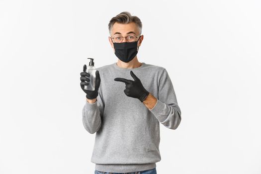 Concept of coronavirus, lifestyle and quarantine. Image of middle-aged man in medical mask and gloves advice to use hand sanitizer, pointing at antiseptic bottle, standing over white background.