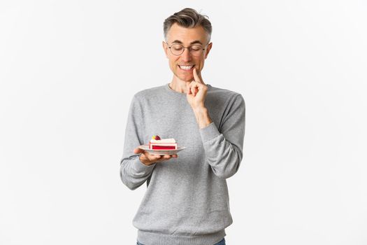 Portrait of handsome middle-aged man in glasses and gray sweater, looking tempted at delicious cake, wanting to eat dessert, standing over white background.