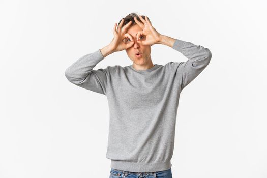 Portrait of funny middle-aged guy making faces, showing hand glasses around eyes and fooling around, standing over white background.