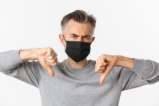 Concept of covid-19, social distancing and quarantine. Close-up of angry and disappointed middle-aged man in black medical mask, frowning upset and showing thumbs-down.