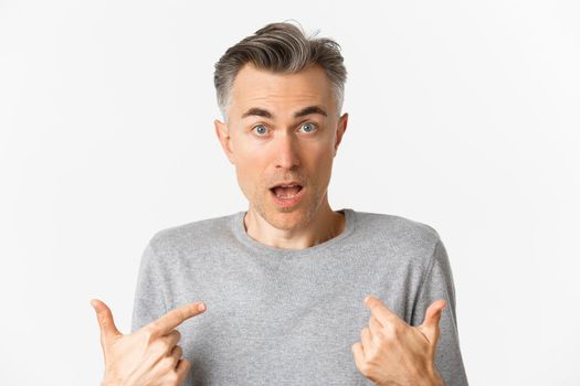 Close-up of confused middle-aged man, pointing at himself and gasping, standing over white background.