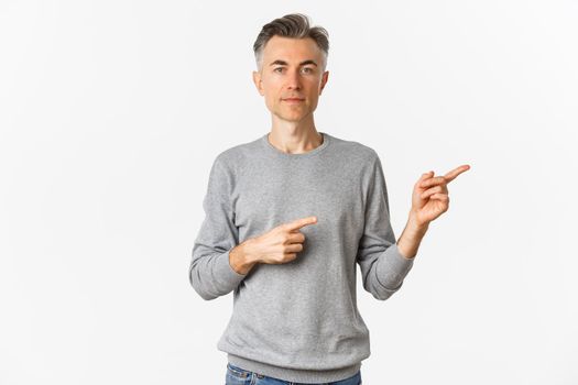 Portrait of attractive middle-aged man in grey sweater, pointing fingers right and looking serious, showing an advertisement, standing over white background.