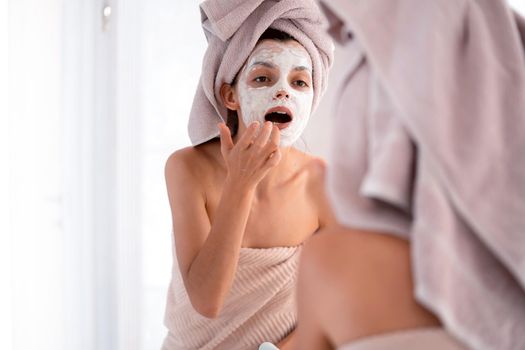 A young girl in a towel on her head and body stands near the mirror in the bathroom and applies a clay mask to her face, the woman takes care of her health and beauty.