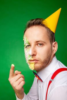 Male in party hat sitting at table and tasting sweet delicious birthday cake while looking up on green background in studio