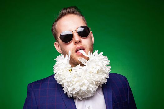 Funny male in suit and sunglasses licking petal of flowers while having fun in studio on green background and looking at camera