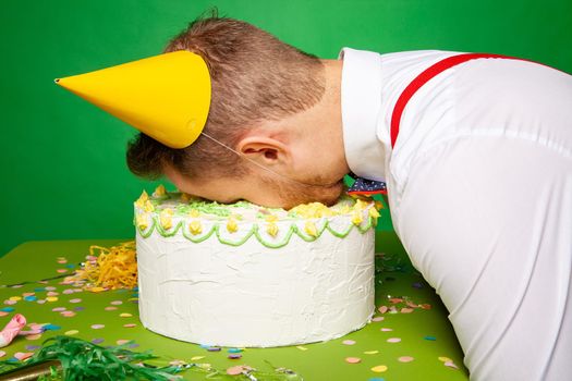 Side view of sleeping male in party hat lying with face in birthday cake on green background in studio