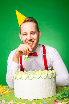 Man in party hat sitting at table with sweet birthday cake and blowing noisemaker on green backwound in studio