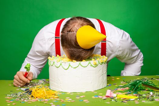 Side view of sleeping male in party hat lying with face in birthday cake on green background in studio