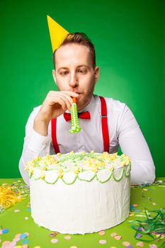 Man in party hat sitting at table with sweet birthday cake and blowing noisemaker on green backwound in studio