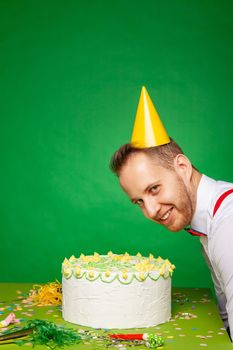 Side view of happy funny male in party hat sitting at table with tasty birthday cake and looking at camera on green background in studio