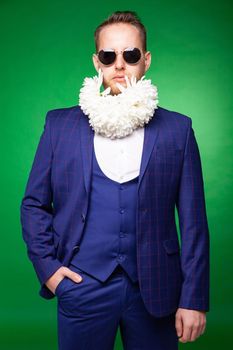Confident male in elegant suit and sunglasses standing with white flowers in beard in studio on green background