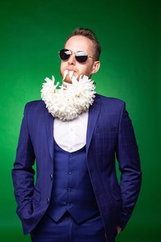 Self confident male in classy suit and flower petals in mouth looking at camera on green background in studio
