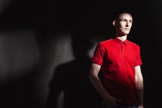 Serious confident young male in bright red shirt looking away while standing under light in dark studio with black wall and shadow