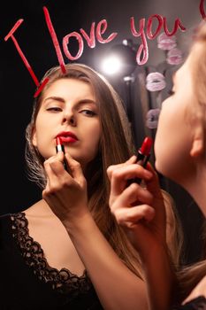 Lovely female looking in mirror with inscription I Love You and applying red lipstick while doing makeup