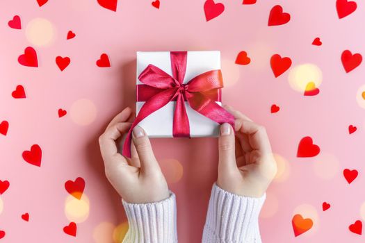 Female Hands in sweater holding a gift in white wrapping paper on pink background with red hearts. St. Valentines Day, love, tenderness, friendship and care concept. Cozy, festive, romantic wallpaper