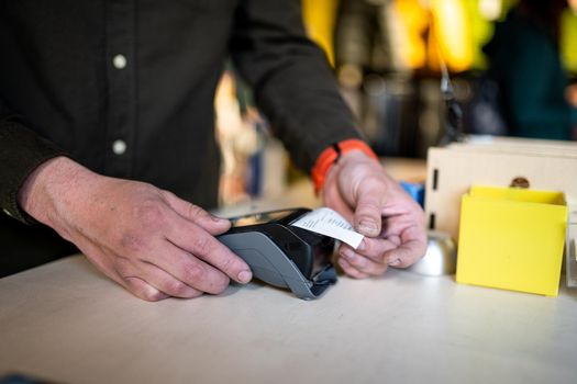 Close-up of the hands of a male salesman cashier holding POS terminal and paper receipt behind the counter of a sports store. Cashless technology and credit card payment concept. Card swipe machine.