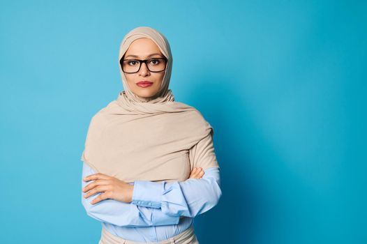 Portrait of serious young arab woman wearing hijab and glasses standing against blue background with crossed arms on chest