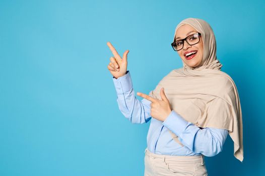 Charming Arab woman in hijab with beautiful smile points her index fingers on blue background with copy space