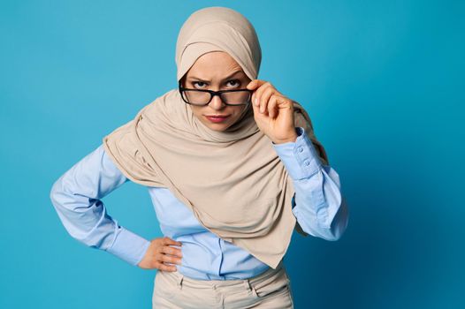 Portrait of a Muslim woman in hijab holding eyeglasses by the temple and looking carefully through them with raised eyebrow, isolated over blue background with copy space