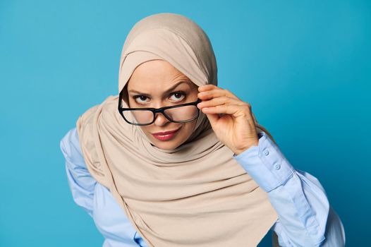 Arab Muslim woman holds eyeglasses by the temple and looks carefully through them with raised eyebrow, isolated over blue background with copy space