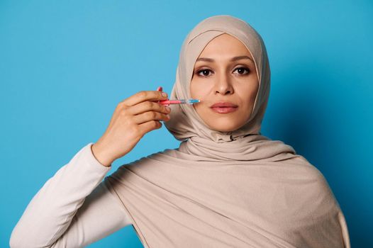 Isolated beauty portrait of a young Arab Muslim woman holding a syringe near her face. Blue background with space for text