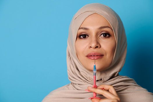 Closeup portrait of beautiful Arab Muslim woman with covered head in hijab holding a syringe with beauty injection near her lips. Lips augmentation concept in injection cosmetology. Blue background, copy space
