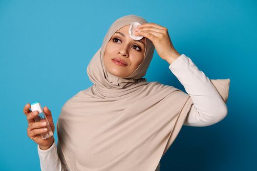 Attractive muslim woman removing makeup from her face using micellar water and cotton pad. Skincare concepts on blue background with copy space