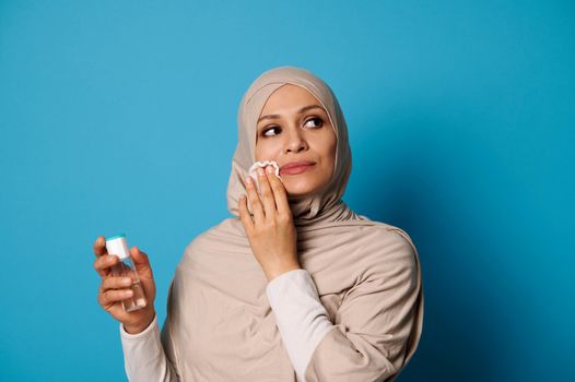 Beautiful Muslim woman with covered head in hijab applying micellar lotion and removing make up from her lips. Make-up removal concepts on blue background with space for text