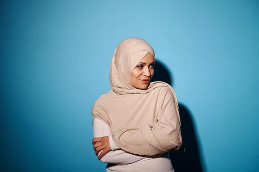 Young woman of oriental appearance in beige hijab, posing over blue background. Copy space. Arabian fashion style