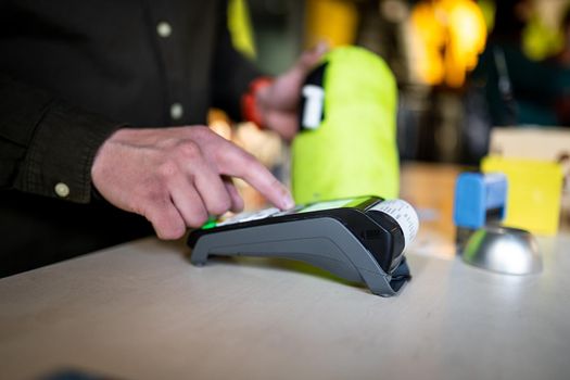 Close-up of the hands of a male salesman cashier holding POS terminal and paper receipt behind the counter of a sports store. Cashless technology and credit card payment concept. Card swipe machine.