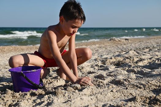 Handsome teenager in red swimming trunks playing on the sandy beach. Adorable child builds sand figures with wet sand against the background of the sea