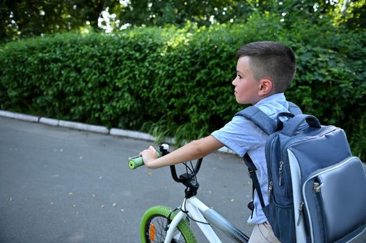 Handsome schoolboy with backpack biking to school on asphalt road.