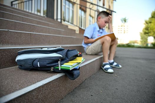 Back to school concept. Upset worried boy with backpack doing his homework. School bag with copy books and school supplies lying on the stairs. Back to school concept